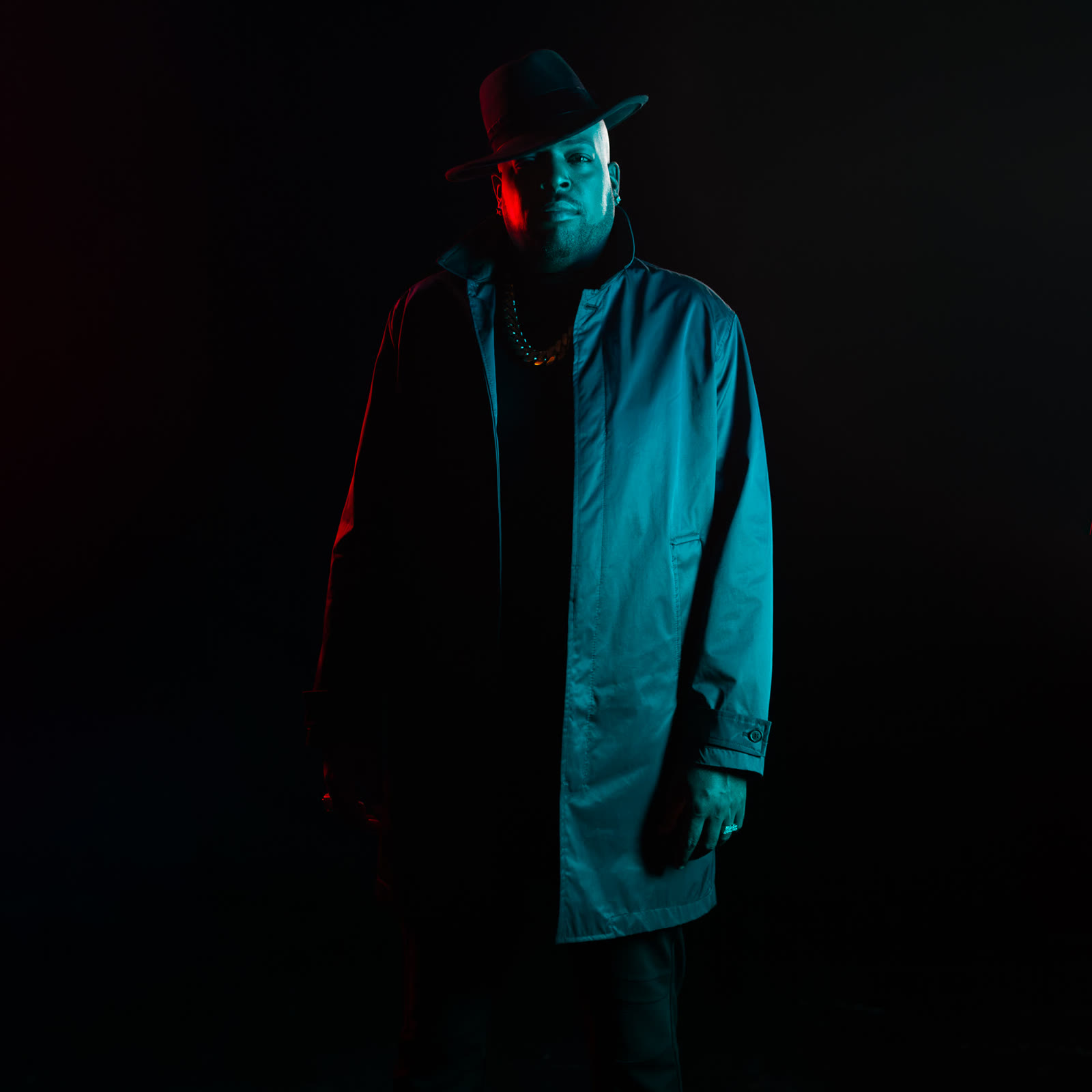 Retrato de cuerpo entero con sombrero y abrigo, entre luz roja y azul sobre fondo negro